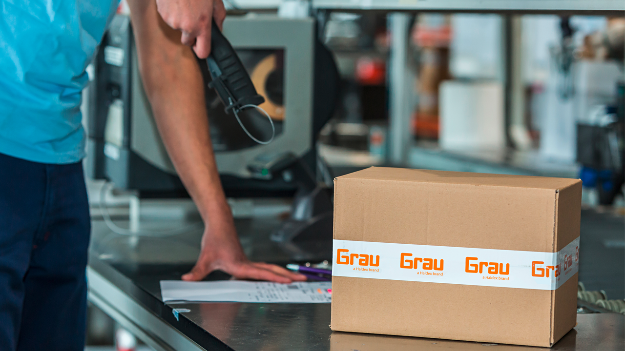 A worker scans documents at a packing station beside a box labeled Grau