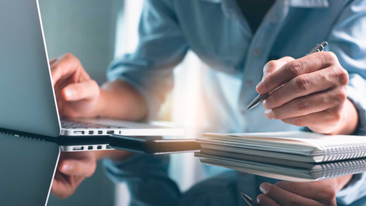 Person working at a desk using a laptop while writing notes in a notebook