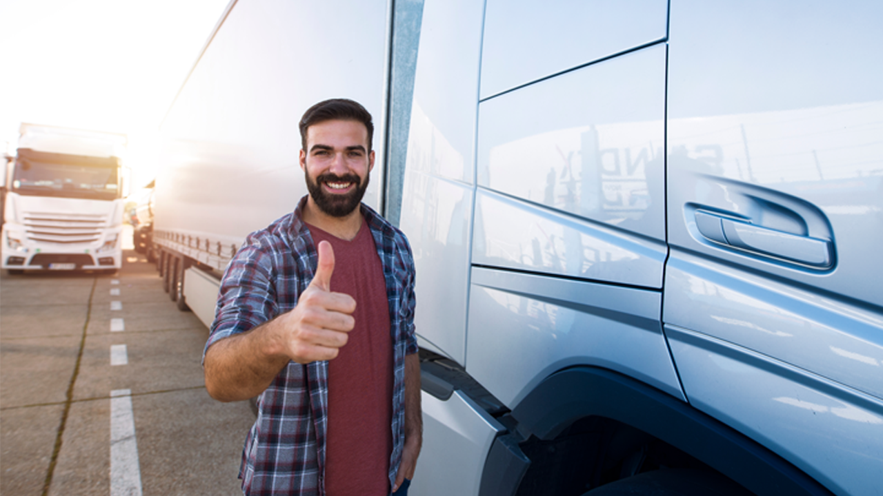 Smiling truck driver stands beside his vehicle giving a thumbs up.