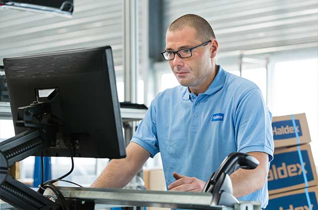 A man in a light blue work shirt uses a desktop computer in an industrial or warehouse setting